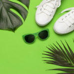 White female fashion sneakers, trendy sunglasses, tropical leaves on green background. Flat lay top view copy space. Women’s shoes, accessories. Stylish white sneakers. Creative summer concept