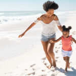 Playful cute little daughter running on beach with young black m