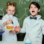 little kids in white coats with chalkboard behind in laboratory