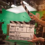 On Film Studio Set Camera Assistant Holds Clapperboard. Green Screen Scene with Talented Cameraman in the Background. Close-up Shot.